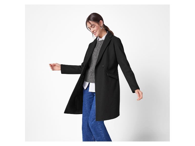 Eine Frau präsentiert einen schwarzen, knielangen Mantel über einem grauen Pullover und blauen Jeans.