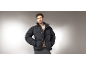 Mann in einer schwarzen Pufferjacke, einem beigen Pullover und einer grauen Hose.