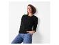 Frau in schwarzem Langarmshirt und blauen Jeans, an eine weiße Wand gelehnt.