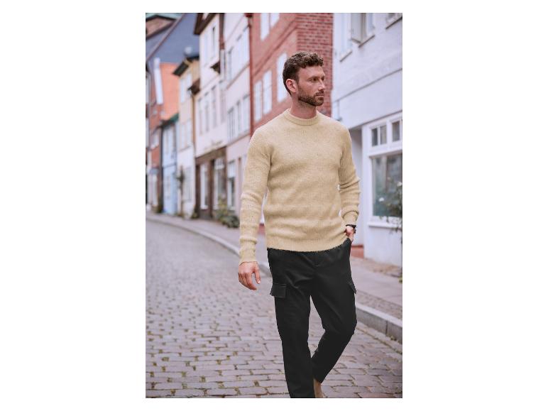 Ein Mann trägt einen beigefarbenen Pullover und eine schwarze Cargohose in einer Stadt.