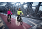 Zwei Personen in reflektierenden Fahrradjacken und Helmen fahren E-Bikes auf einer Brücke.