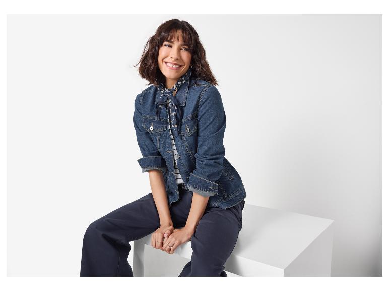 Lächelnde Frau in Jeansjacke, Halstuch und marineblauer Hose, sitzt auf einem weißen Würfel.