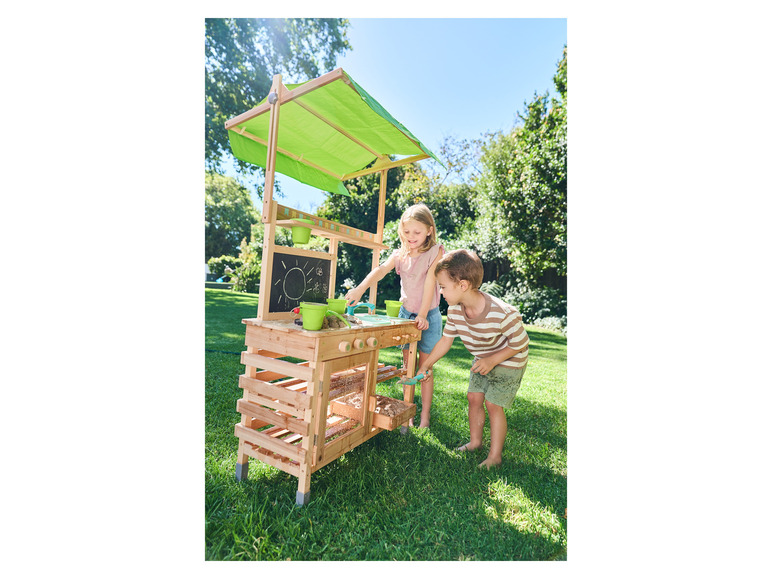 Zwei Kinder spielen im Garten mit einer hölzernen Spielküche mit grünem Sonnendach.