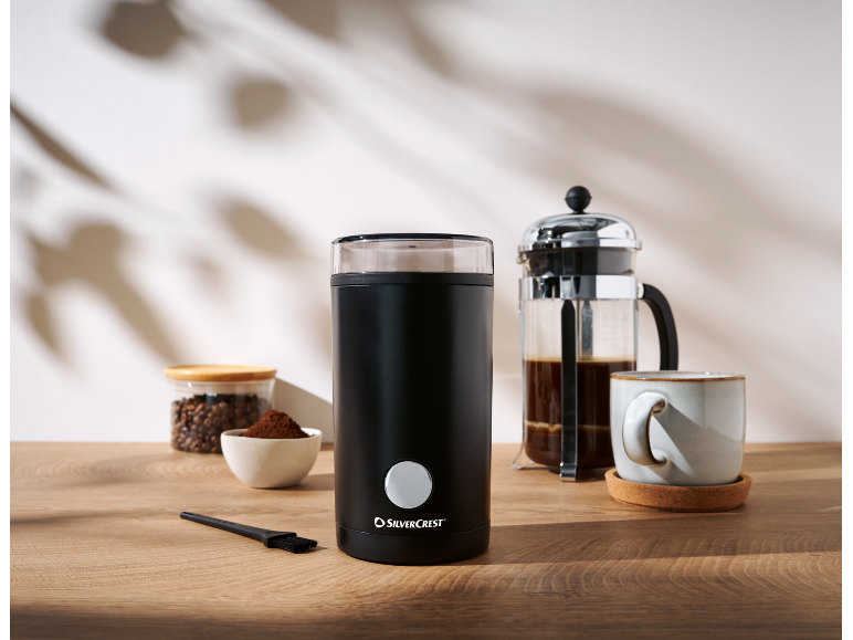 Schwarze elektrische Kaffeemühle, Kaffeebohnen, gemahlener Kaffee und eine French Press auf Holztisch.