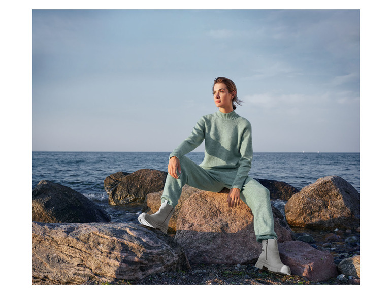 Eine Frau trägt einen grünen Pullover und eine Hose mit weißen Stiefeln und sitzt auf Felsen am Meer.