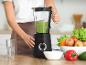 Eine Frau bereitet einen grünen Smoothie in einem Mixer zu, mit frischem Obst und Gemüse auf dem Tisch.