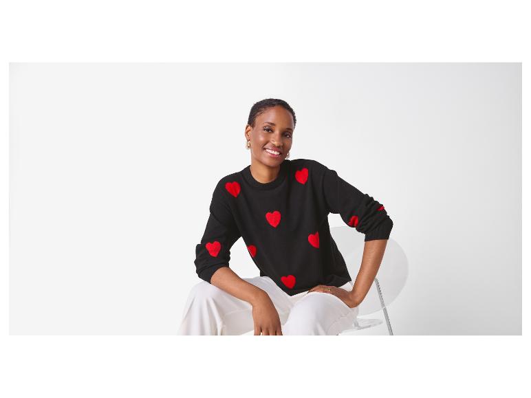 Frau in schwarzem Pullover mit Herzmuster und weißer Hose, auf einem Stuhl sitzend.