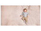 Ein Baby liegt auf einer rosafarbenen Decke, trägt einen grauen Pullover mit Punkten und eine beige Hose.