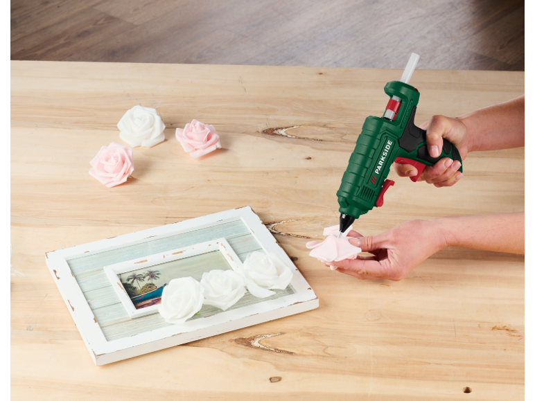 Person verwendet Heißklebepistole, um Rosen an einem Bilderrahmen zu befestigen.