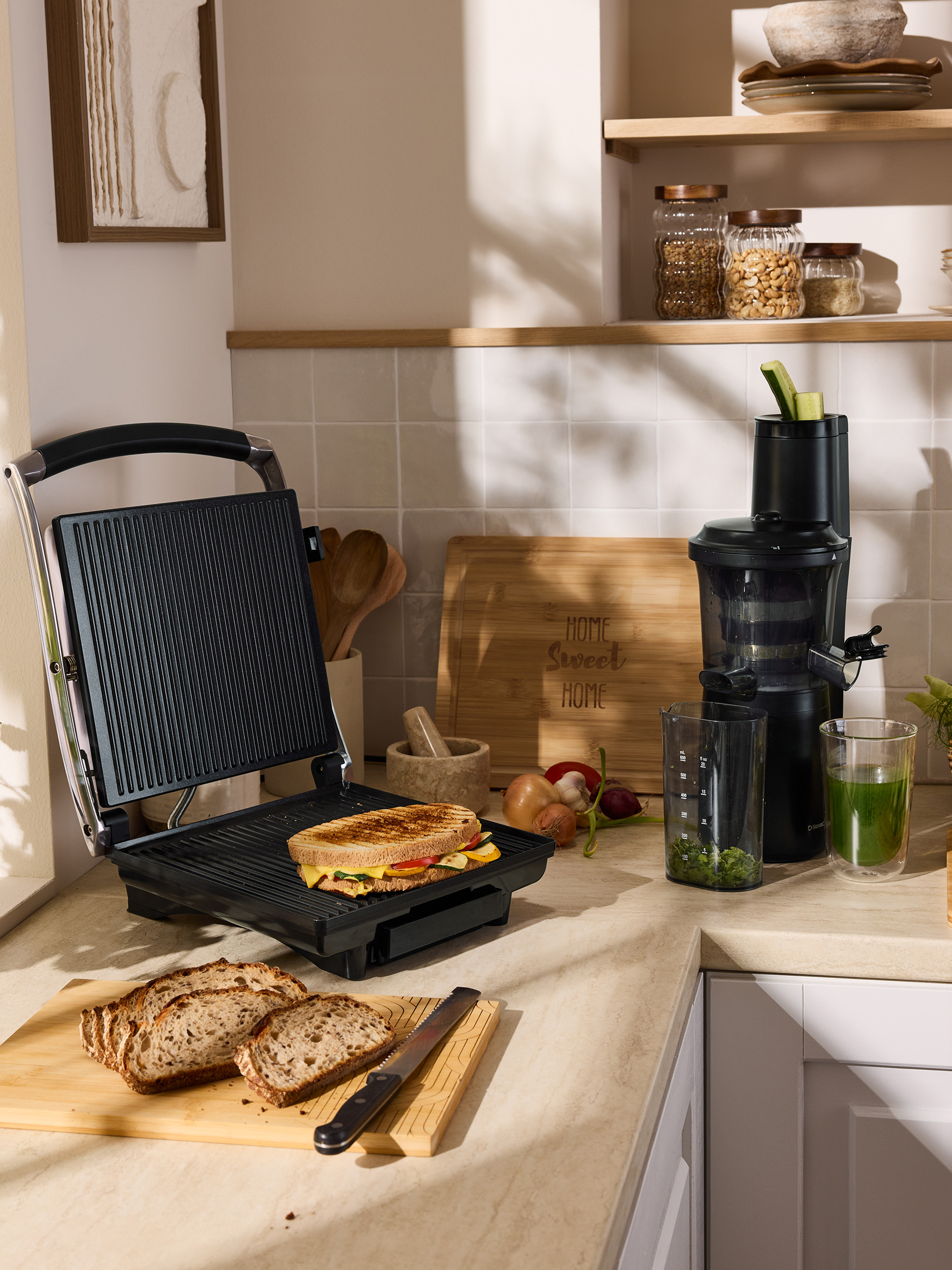 Ein gegrilltes Sandwich auf einem Sandwichmaker, geschnittenes Brot und ein Entsafter auf einer Küchentheke.