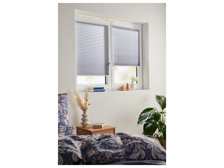 Ein Schlafzimmer mit einem Bett, einem Nachttisch und einem Fenster mit grauen Plissee-Vorhängen.