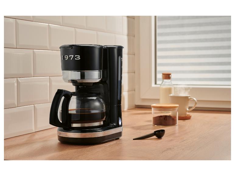 Schwarze Kaffeemaschine mit der Aufschrift „1973“ auf einer Küchenarbeitsplatte mit Frühstückszubehör.