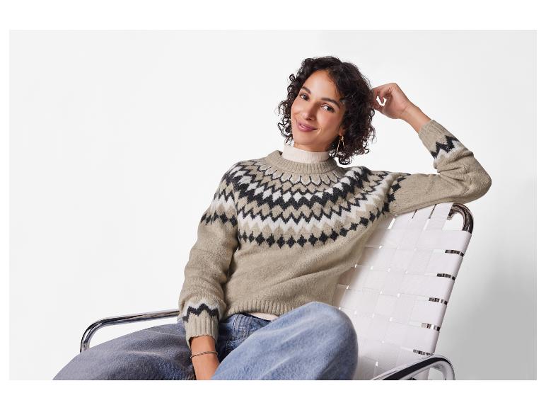 Lächelnde Frau in beigem gemustertem Pullover, Jeans und goldenen Ohrringen, auf einem Stuhl sitzend.