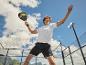 Mann in Sportkleidung spielt Padel mit Schläger und Ball auf einem Außenplatz.