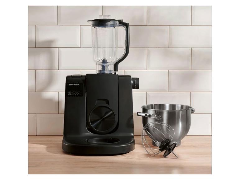 Schwarze Küchenmaschine mit Mixer, Schüssel und Schneebesen auf Holztisch.