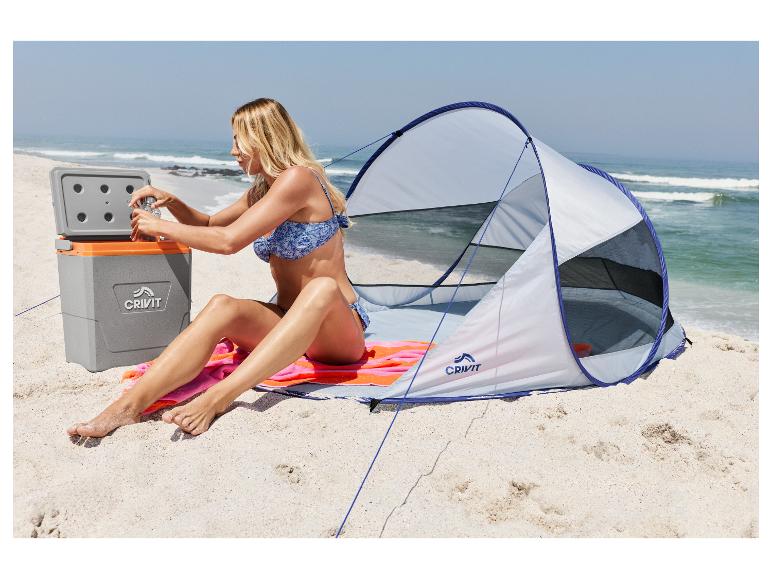 Frau am Strand sitzt neben Zelt und Kühlbox, öffnet eine Flasche.