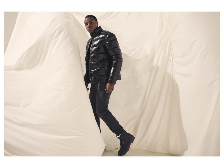 Mann in glänzender schwarzer Pufferjacke und dunklen Jeans vor weißem Stoff.