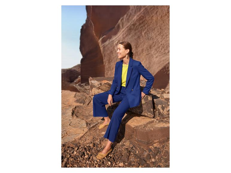 Frau in blauem Anzug und gelbem Oberteil, sitzt auf Felsen in einer Wüstenlandschaft.