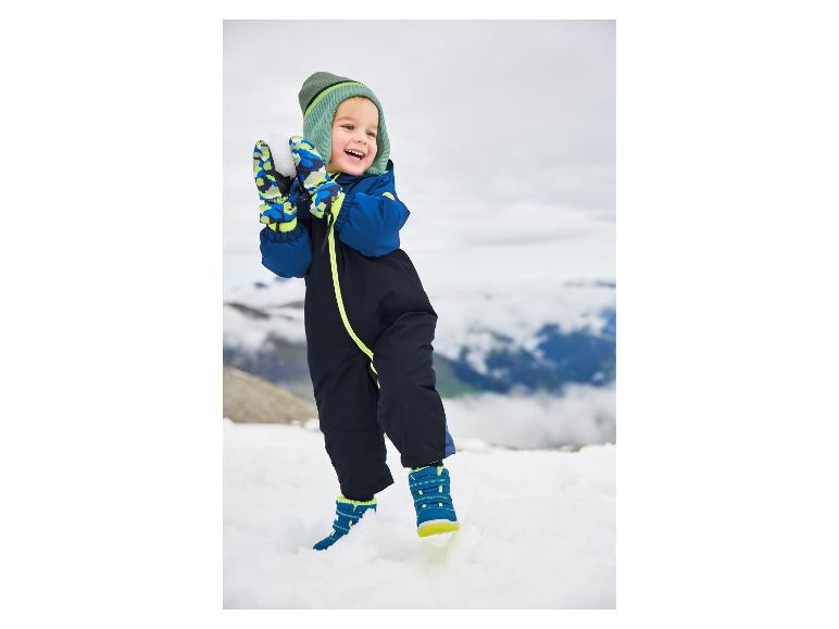 Glückliches Kleinkind in blauem Schneeanzug und grüner Mütze spielt im Schnee.