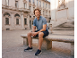 Mann in blauem Poloshirt und marineblauen Shorts sitzt auf einer Steinbank auf einem Stadtplatz.