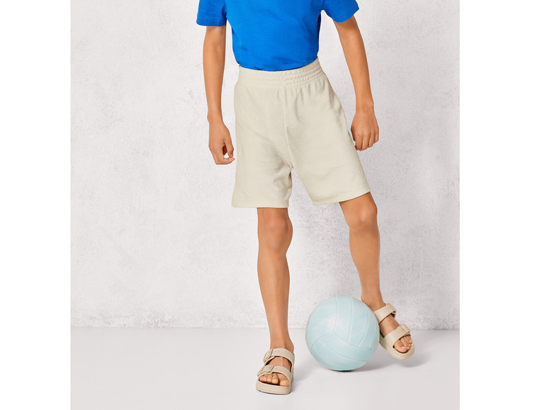 Ein Junge trägt beige Shorts, Sandalen und ein blaues T-Shirt, während er mit einem Volleyball spielt.