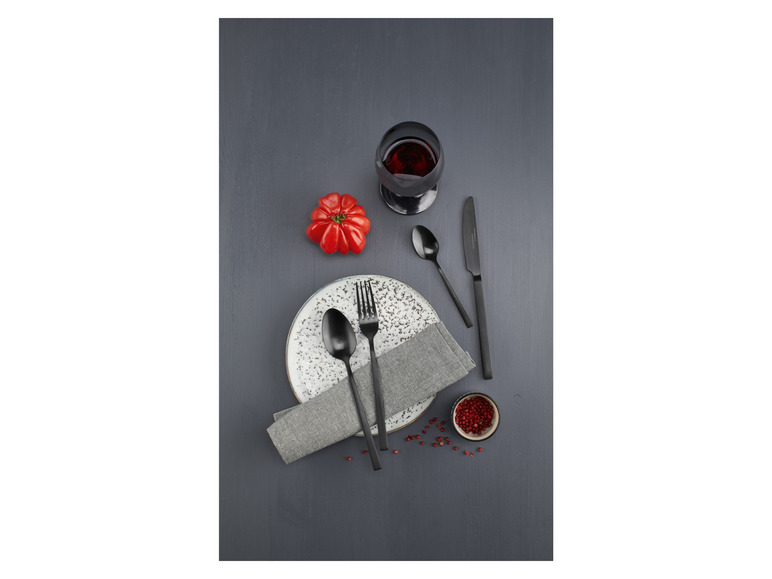 Schwarzes Besteck, ein Teller, eine Serviette, ein Glas Rotwein und Pfefferkörner auf einem dunklen Holztisch.