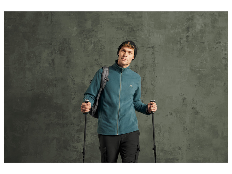 Ein Mann in einem grünen Fleece-Pullover mit Rucksack und Wanderstöcken.
