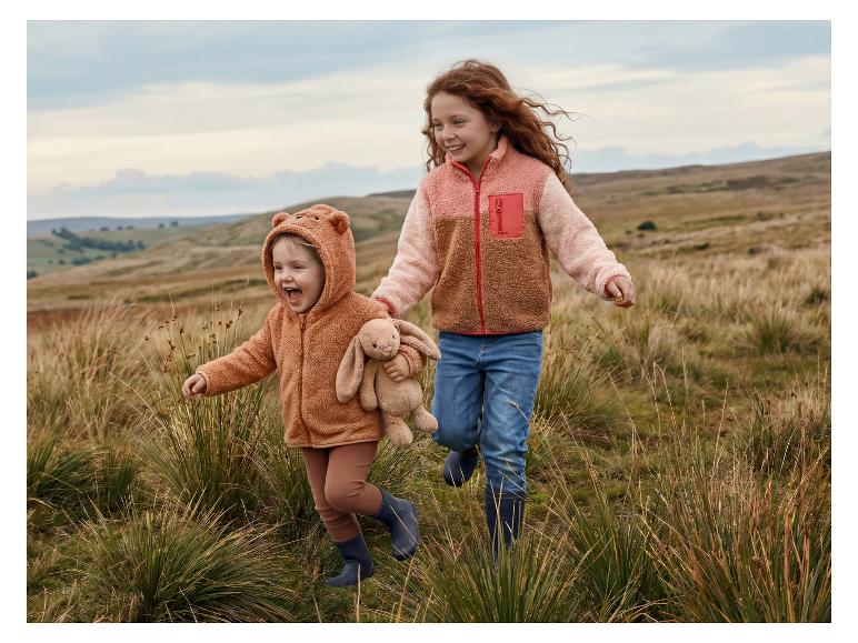 Zwei glückliche Kinder in Fleecejacken und Gummistiefeln rennen durch ein Feld.