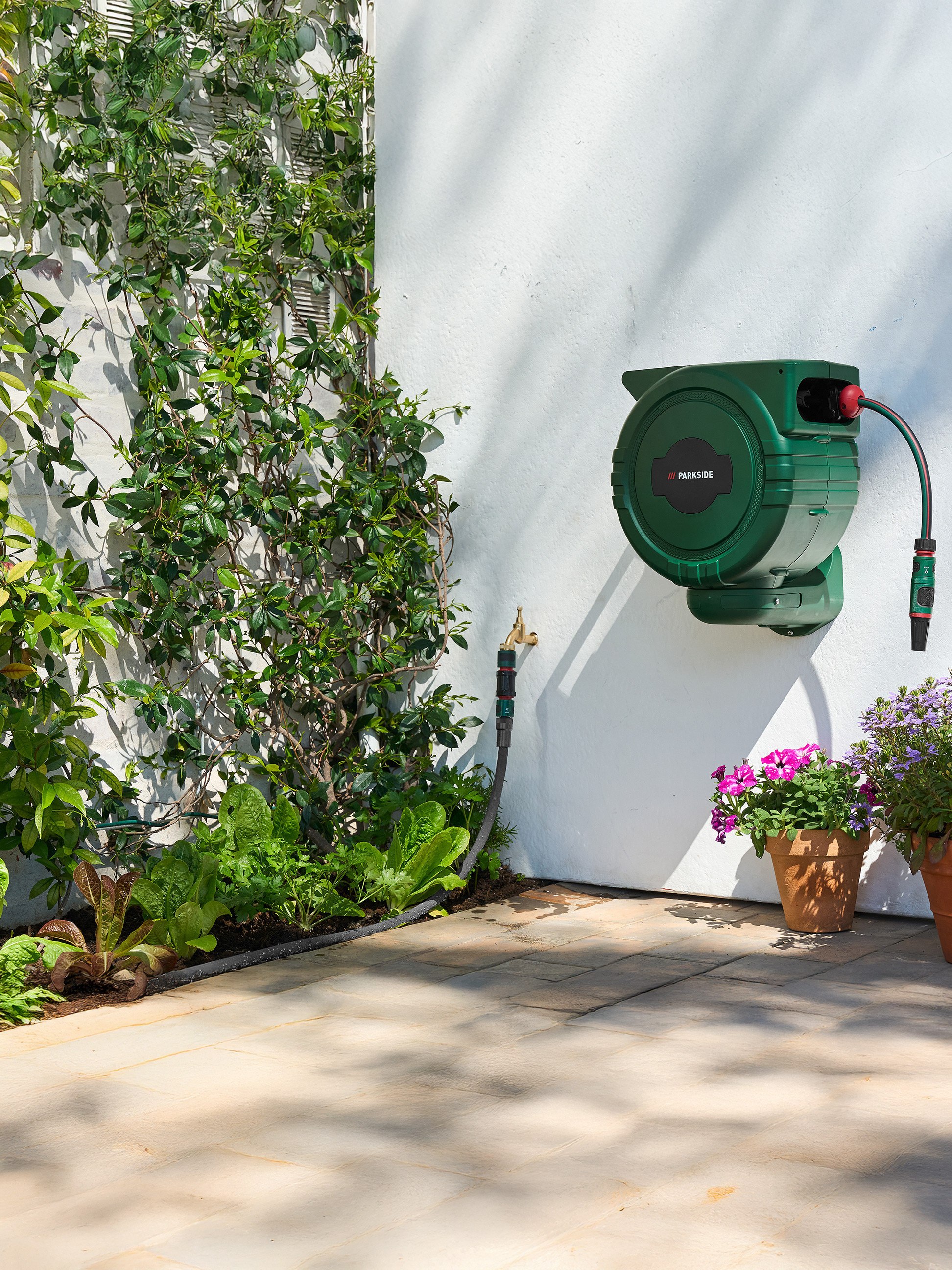 Grüner PARKSIDE Gartenschlauchaufroller an einer weißen Wand mit Kletterpflanzen und Gemüsebeet.