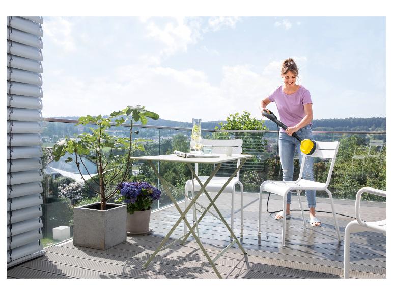 Frau reinigt Balkonmöbel mit einem Hochdruckreiniger auf einem Balkon mit Aussicht.