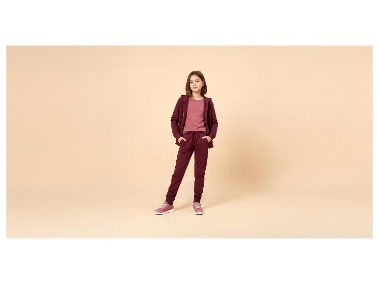 Mädchen in bordeauxfarbenem Trainingsanzug und rosa T-Shirt, posiert mit Händen in den Taschen.