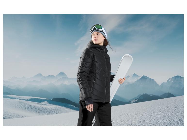 Frau in einer schwarzen Jacke mit Skibrille hält weiße Ski vor einer Bergkulisse.