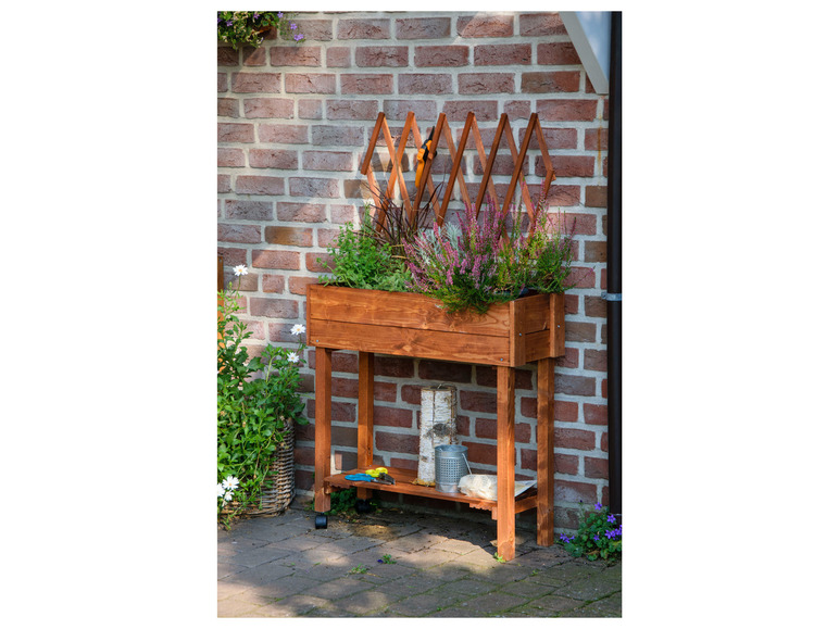 Ein Blumenkasten aus Holz mit Blumen auf Rädern vor einer Backsteinwand.