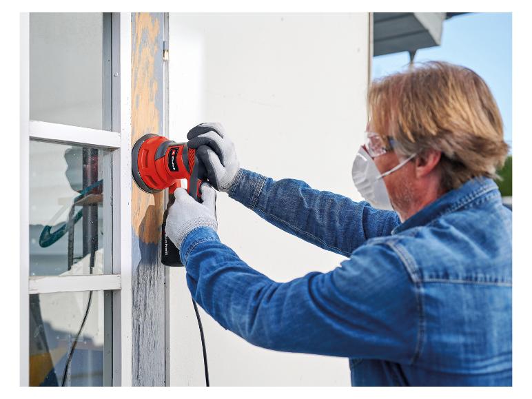 Mann schleift Fensterrahmen mit Elektroschleifer, trägt Schutzbrille und Maske.
