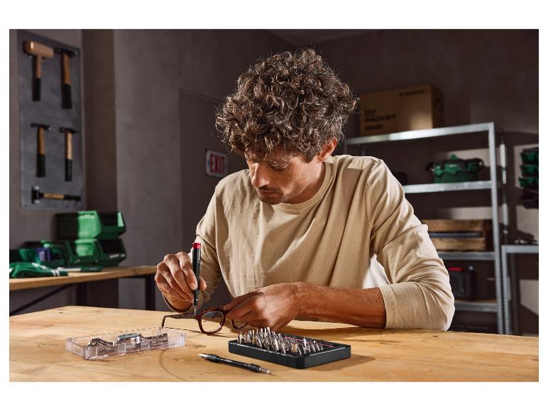 Mann repariert Brille mit Präzisionsschraubendreher und Bit-Set auf einer Holzwerkbank.
