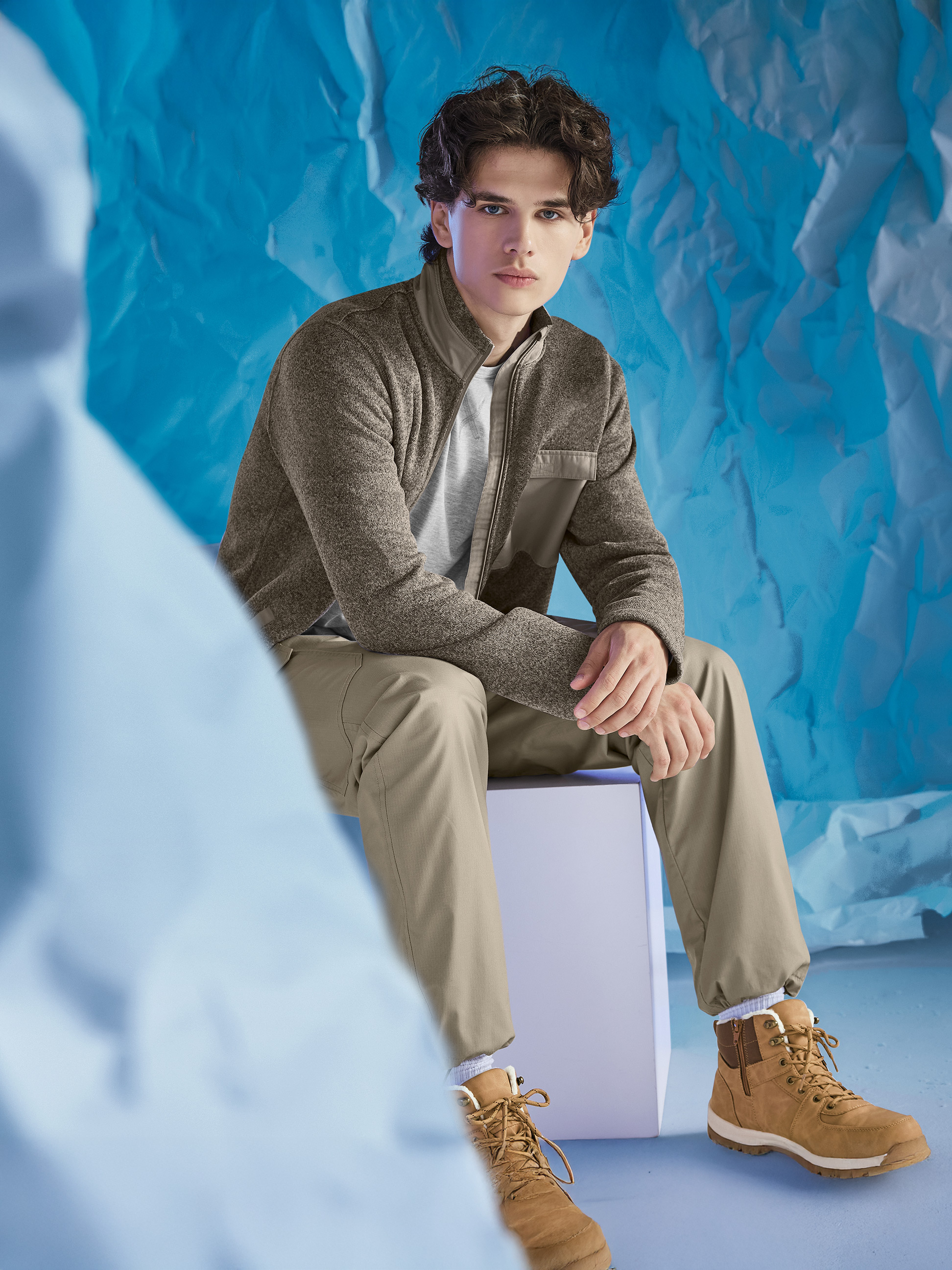 Ein Mann trägt eine braune Jacke, ein graues T-Shirt und khakifarbene Hosen, sitzend vor blauem Hintergrund.