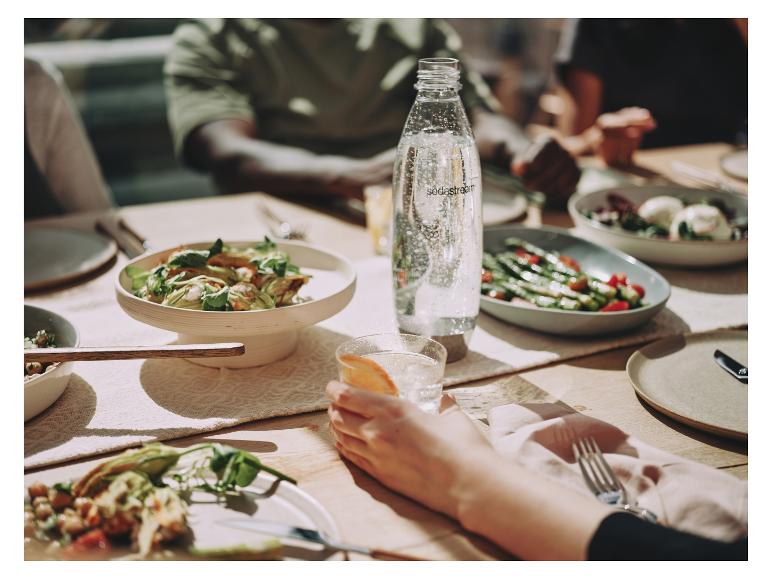 Eine Sodastream-Flasche mit Sprudelwasser auf einem Tisch mit Essen und Menschen.