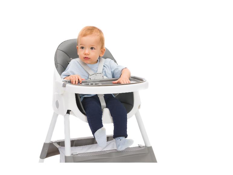 Baby sitzt in einem Hochstuhl mit grauem Tablett vor weißem Hintergrund.