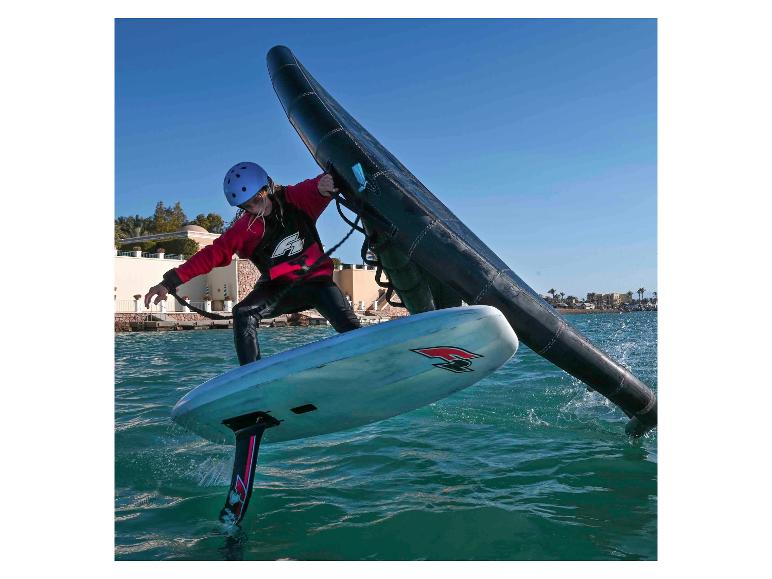 Frau in Helm und Neoprenanzug beim Windfoiling mit Flügel und Foilboard.