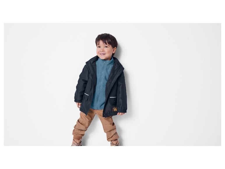 Junge in dunkelblauer Jacke mit türkisfarbenem Fleece und beiger Hose, stehend und lächelnd.