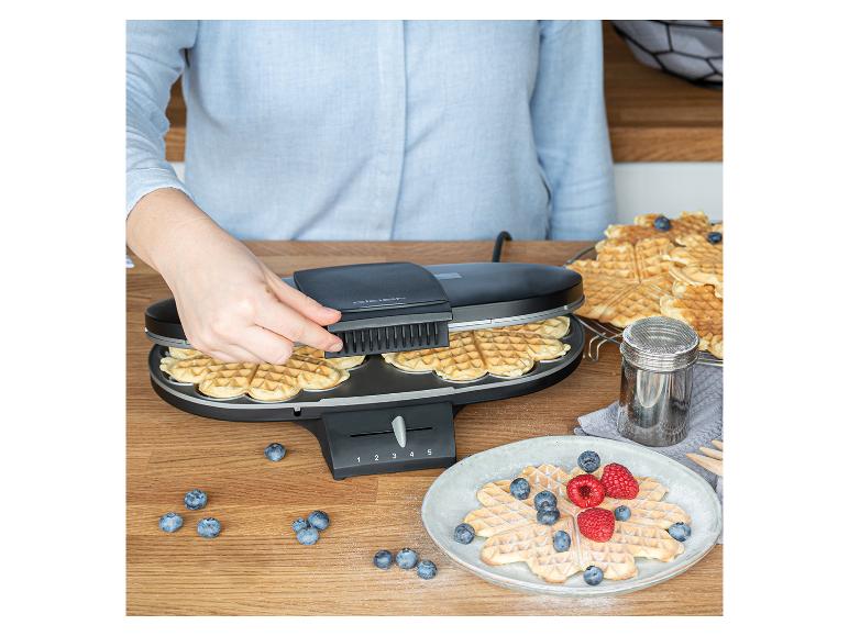 Person macht Waffeln in einem Waffeleisen, mit Waffeln und Beeren auf dem Tisch.
