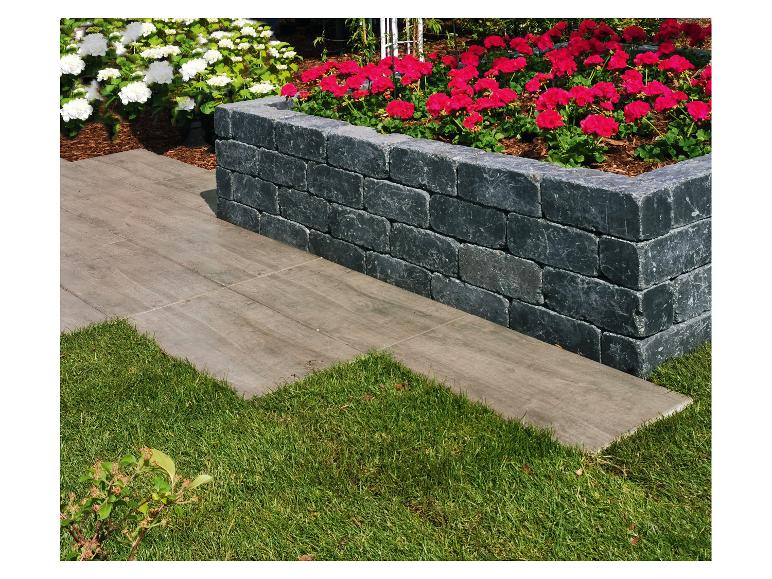 Graues Steinblock-Pflanzgefäß mit roten Geranien und weißen Hortensien, umrandet von einem gefliesten Weg und Rasen.