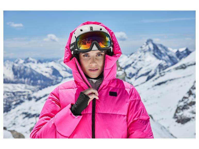 Frau in pinker Skijacke und Helm mit Skibrille vor verschneiter Bergkulisse.