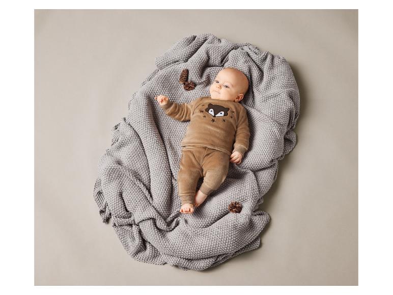 Ein Baby liegt auf einer grauen Decke in einem braunen Strampler mit einem Fuchs-Motiv.