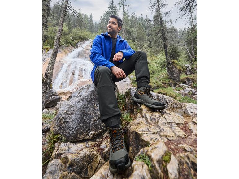 Mann in blauer Jacke und schwarzen Hosen in der Nähe eines Wasserfalls, mit schwarzen Wanderschuhen.