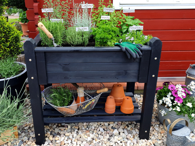 Ein schwarzer Pflanzenkasten mit Kräutern wie Rosmarin, Thymian und Oregano, sowie Terrakotta-Töpfen und einer Gießkanne.