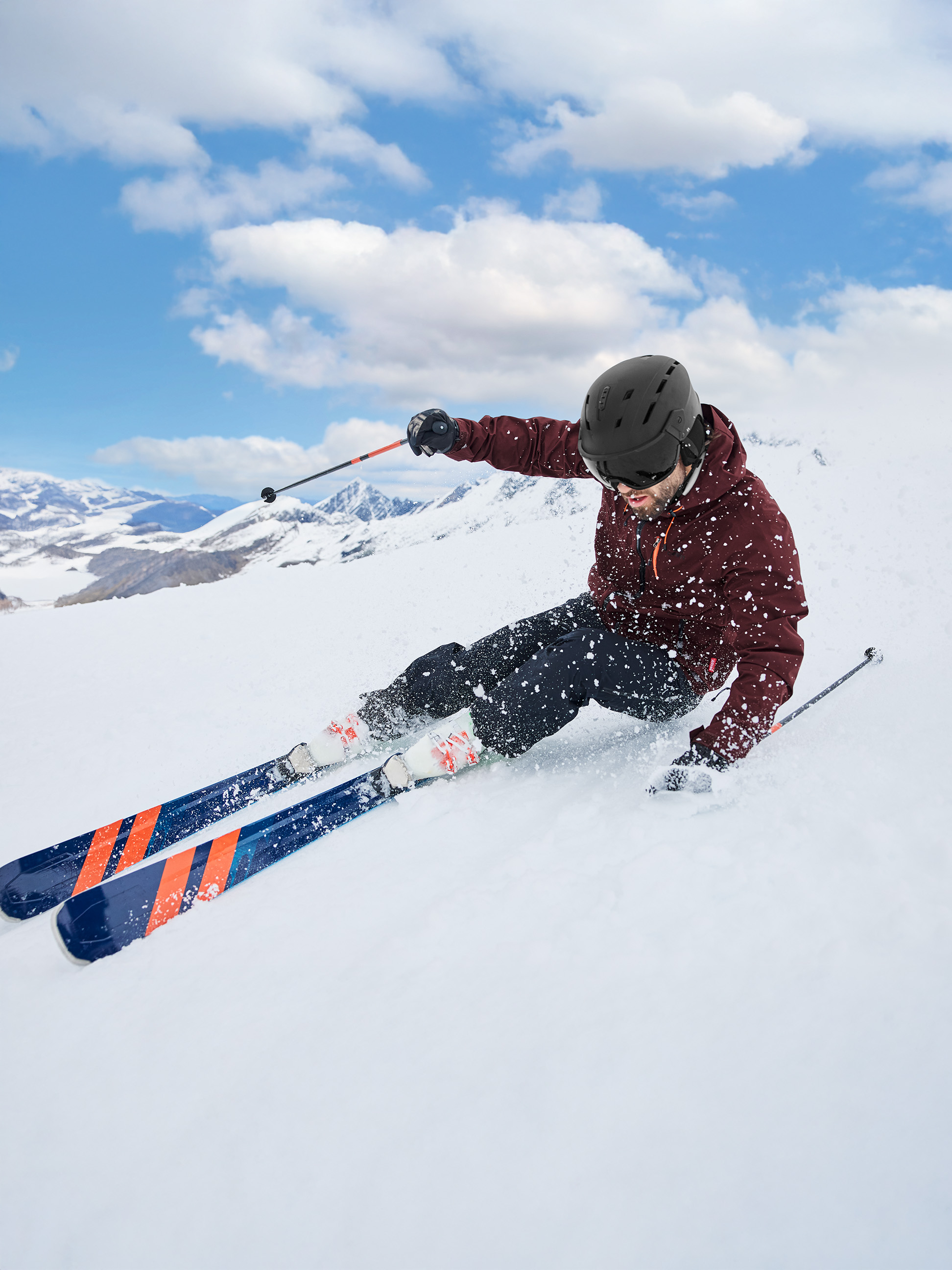 Ein männlicher Skifahrer in einer bordeauxroten Jacke und schwarzem Helm fährt einen verschneiten Berg hinunter.