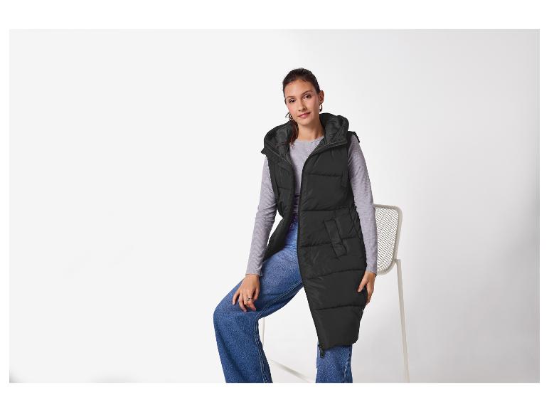 Frau in schwarzer Kapuzen-Steppweste, grauem Shirt und Jeans, auf einem Stuhl sitzend.