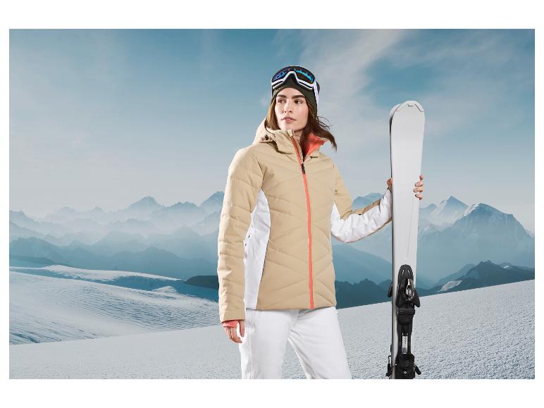 Eine Frau in einer beige Skijacke und einer weißen Skihose hält Skier vor einer Bergkulisse.