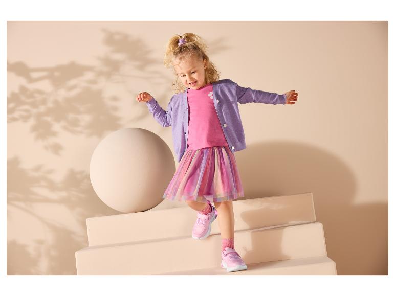 Lächelndes Mädchen in rosa Top, lila Strickjacke und buntem Rock, trägt rosa Sneaker.